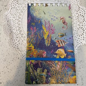 May include: A spiral-bound notebook with a vibrant underwater scene featuring colorful fish, coral, and a seahorse. The cover has a blue elastic band. The notebook is resting on a white lace doily.