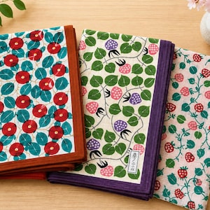 May include: Three folded rectangular cloths featuring distinct botanical patterns. The left cloth has red flowers on teal leaves with a brown border. The center cloth displays pink and purple berries on green leaves with a purple border and a small white label with Japanese text. The right cloth shows red strawberries on green vines against a light pink background.