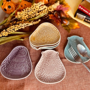 May include: Four ceramic dishes featuring animal designs. A purple owl, a pink hedgehog, a blue bear, and a yellow fox. The dishes are displayed on a brown surface with autumn leaves and pumpkins.