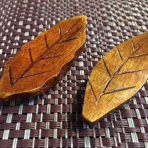 Puede incluir: Dos posavasos de madera en forma de hoja con un diseño tallado. Los posavasos son de color marrón y tienen un acabado de madera natural.