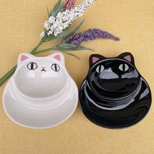 Könnte beinhalten: Zwei katzenförmige Keramikschalen, eine weiße und eine schwarze, nebeneinander. Jede Schale hat ein Katzengesicht-Design mit rosa Akzenten an Ohren und Nase. Die Schalen stehen auf einer gelben Oberfläche, mit dekorativen lila und weißen Blumen.