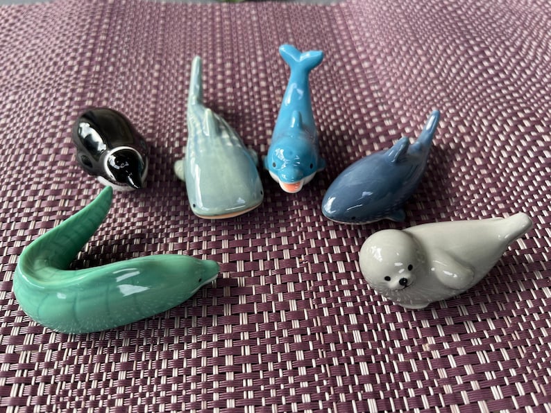 May include: A collection of ceramic marine animal figurines, including a black penguin, a gray whale, a blue dolphin, a dark blue dolphin, a green eel, and a gray seal. The figurines are arranged on a purple woven surface.