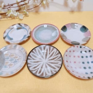 May include: Six small ceramic plates with various designs. The plates are white with different patterns in shades of blue, green, brown, pink, and black. The plates are arranged in a circle on a yellow surface.