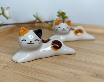 May include: Two ceramic cat chopsticks rests. The cats are white with orange and black markings. They are laying on their bellies with their paws outstretched.