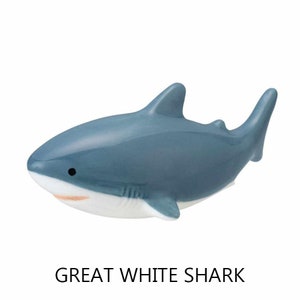 May include: A ceramic figurine of a great white shark. The shark is primarily blue-gray with a white underbelly and a small black eye. The text "GREAT WHITE SHARK" is printed in black below the figurine.