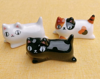 May include: Three ceramic cat figurines, one white, one black, and one calico. The cats are all in a lying down position with their eyes closed.