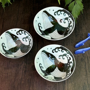 May include: Three white ceramic bowls with black and white whale designs. The bowls have a blue and white wave pattern around the whale. Two blue whale-shaped serving utensils are on a wooden surface.
