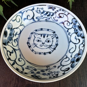 May include: A white ceramic bowl with a blue and white cat design. The bowl features a cat face in the center, surrounded by a floral pattern. There are two cats on the rim of the bowl, one with a butterfly.
