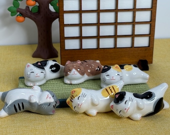 May include: Set of six ceramic cat chopsticks rests. The cats are in various sleeping positions and are painted in white, black, gray, and brown with yellow accents.