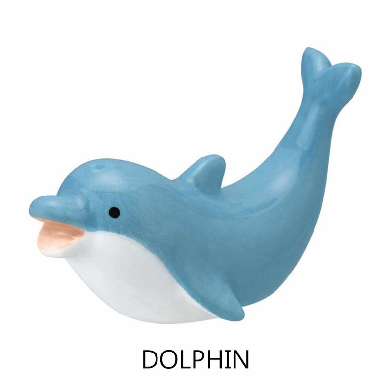 May include: A light blue dolphin figurine with a white belly and open mouth. The figurine has a curved body and a raised tail. The word "DOLPHIN" is printed in black below the figurine.