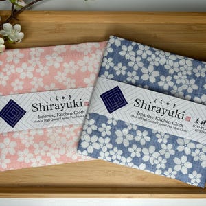 Puede incluir: Dos paños de cocina japoneses con un patrón floral. Los paños están hechos de tejido de malla fina en capas y están teñidos con la técnica Kyo-Yuzen. Los paños son rosa y azul con flores blancas.