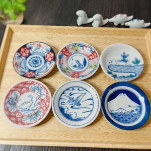 Peut inclure: Six petites assiettes rondes en céramique avec des motifs floraux et géométriques bleus et rouges. Chaque assiette présente une image différente, notamment un dinosaure, un oiseau, une montagne et une vague.