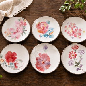 Peut inclure: Un ensemble de six assiettes rondes en céramique blanche, chacune ornée d'une illustration florale unique de style aquarelle. Les motifs présentent diverses fleurs roses, rouges et violettes avec des feuilles vertes, disposées en deux rangées.