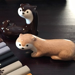 May include: Two ceramic otter chopsticks holders. One is brown and white, the other is light brown. They are both lying on a dark wooden surface.