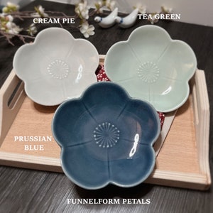 May include: Three small ceramic dishes with a flower-shaped design. The dishes are in cream, light green, and blue colors. The text "FUNNELFORM PETALS" is printed below the dishes. The dishes are labeled "CREAM PIE", "TEA GREEN", and "PRUSSIAN BLUE".