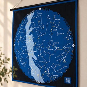 May include: A rectangular wall hanging featuring a circular star map on a dark blue and black background. The map displays constellations with white lines and yellow stars, labeled with Japanese text. It is suspended from a black rod.