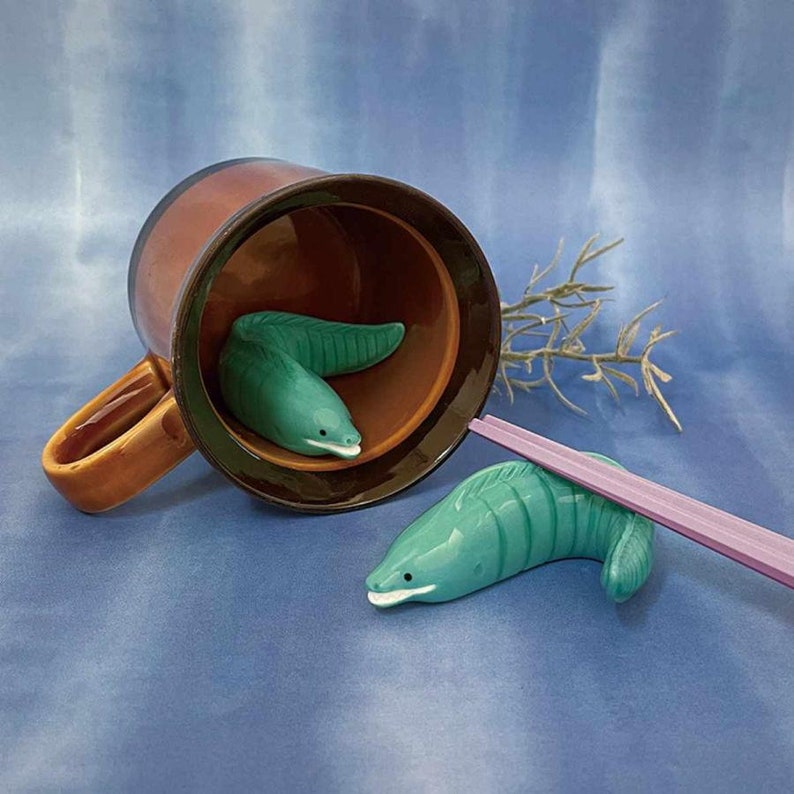 May include: A brown ceramic mug with a tan handle, containing two teal eel-shaped ceramic chopsticks rests. A third eel-shaped chopstick rest is next to the mug, with a pair of pink chopsticks resting on it. The background is blue.