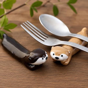 May include: Two ceramic otter-shaped chopstick rests, one dark brown and one light tan, designed to hold cutlery. A stainless steel fork rests across the tan otter, while the dark brown otter lies empty. Both figures feature white faces with black eyes and noses.