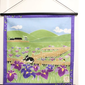 May include: A colorful wall hanging with a black cat sitting in a field of purple irises. The background features a green hill and a blue sky with white clouds. The text on the wall hanging reads "対馬の  故郷".