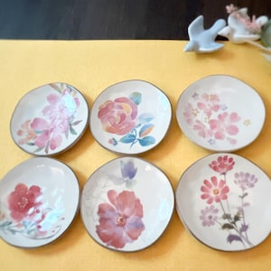 Peut inclure: Six petites assiettes en céramique blanche avec des motifs floraux. Chaque assiette présente un motif floral différent de style aquarelle dans des tons de rose, bleu et vert.