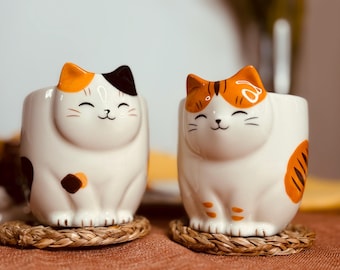 May include: Two ceramic cat mugs with orange, black, and white designs. The mugs are sitting on woven coasters.