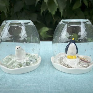 May include: Two white ceramic snow globe-style trinket dishes. One features a white polar bear figurine and the other features a blue and white penguin figurine. Both dishes have a white base and a clear glass dome.