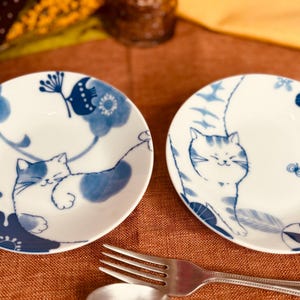 May include: Two white ceramic plates with blue and white floral designs and a cat illustration. The plates are on a brown surface.