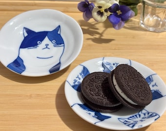 May include: Two small, white ceramic plates with blue cat illustrations. One plate holds two chocolate sandwich cookies. The other plate features a single cat face. The plates are on a wooden surface.