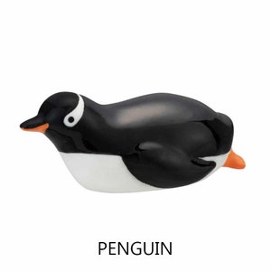 May include: A ceramic penguin figurine with a glossy black body, white belly, and orange beak and feet. The penguin has a white eye marking and is lying down. The word "PENGUIN" is printed in black below the figurine.