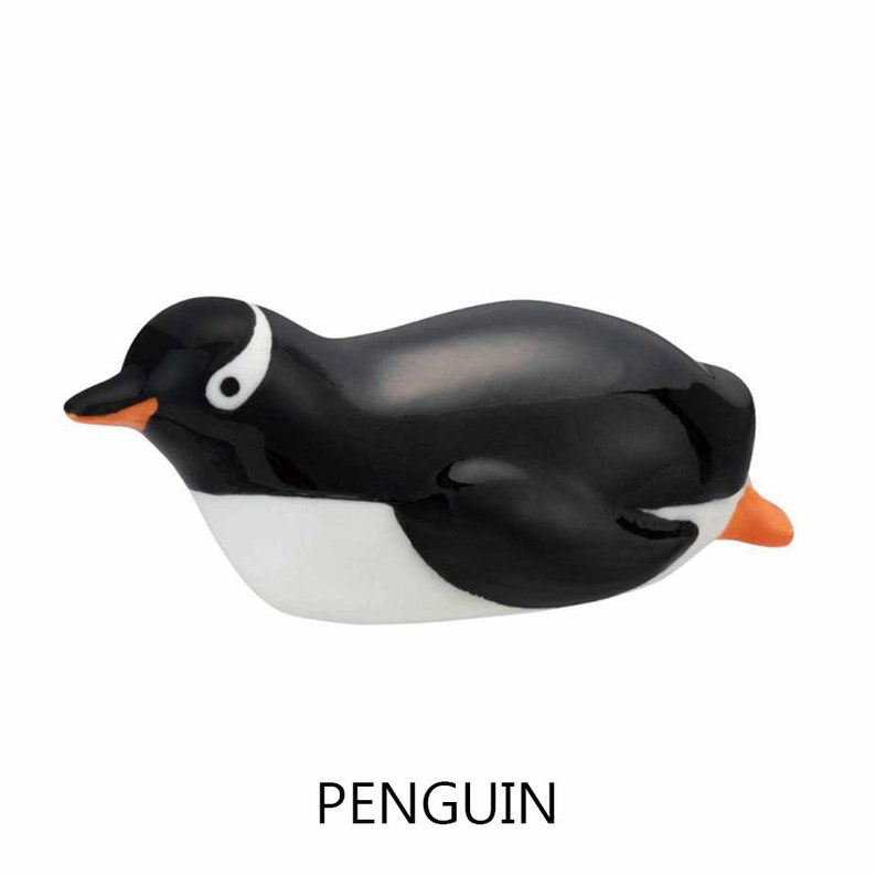 May include: A ceramic penguin figurine with a glossy black body, white belly, and orange beak and feet. The penguin has a white eye marking and is lying down. The word "PENGUIN" is printed in black below the figurine.