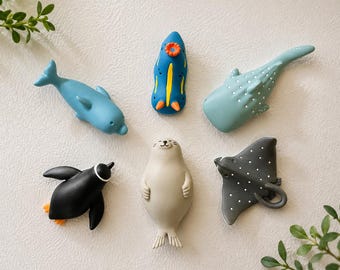 May include: A collection of six small animal-shaped figures including a blue dolphin, a blue and yellow sea slug, a spotted whale shark, a black and white penguin, a beige seal, and a grey spotted stingray, all arranged in a circular formation.