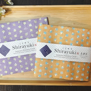 Może przedstawiać: Dwa japońskie ściereczki kuchenne z wzorem grzybów. Ściereczki są fioletowe i pomarańczowe z tekstem "Shirayuki Japanese Kitchen Cloth Made of High Quality Layered Fine Mesh Cloth" i "Shirayuki Japanese Kitchen Cloth Made of High Quality Layered Fine Mesh Cloth KYO-YUŽIN DYEING" na etykietach.