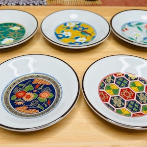 May include: Five small white ceramic plates with black rims. Each plate features a different colorful floral or geometric design.