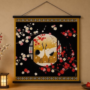 May include: A Japanese-style wall hanging depicting two cranes in a circular window against a golden backdrop. The black fabric is decorated with red and white blossoms. The artwork is framed with a black and gold border and is suspended by a black rod.