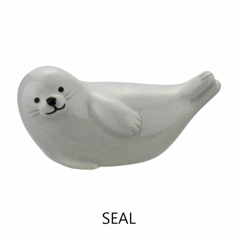 May include: A ceramic seal figurine in a reclining pose. The seal is a light gray color with a black nose, eyes, and whiskers. The word "SEAL" is printed in black below the figurine.