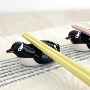 May include: Black and white ceramic penguin chopstick rests with orange beaks and white eyes. Two pairs of chopsticks rest on the penguin-shaped holders. One pair is yellow, and the other is pink. The penguin rests are on a striped cloth.