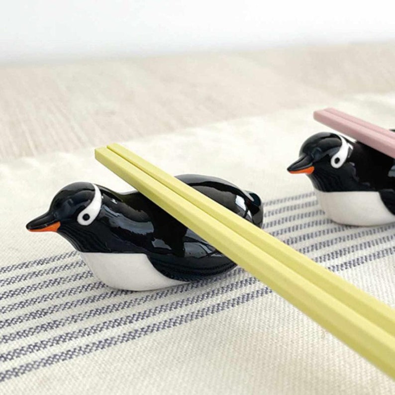 May include: Black and white ceramic penguin chopstick rests with orange beaks and white eyes. Two pairs of chopsticks rest on the penguin-shaped holders. One pair is yellow, and the other is pink. The penguin rests are on a striped cloth.