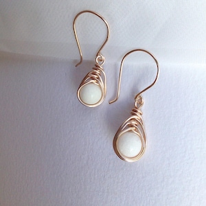 May include: A pair of gold-coloured wire-wrapped earrings. Each earring has a teardrop shape with a white, rounded stone at the centre. The earrings have a simple hook closure and are shown against a white background.