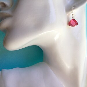 May include: A single earring with a triangular, ruby-colored gemstone is displayed. The gemstone is suspended from a silver hook. The earring is shown against a white background and a light blue backdrop.