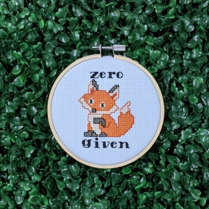 May include: A cross stitch pattern of a fox with the text "zero given" on a white background. The pattern is framed in a wooden hoop.