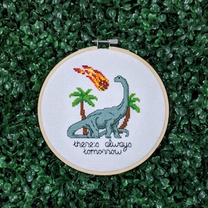 May include: A cross stitch pattern featuring a green dinosaur with a meteor falling from the sky. The text "There's always tomorrow" is stitched below the dinosaur.