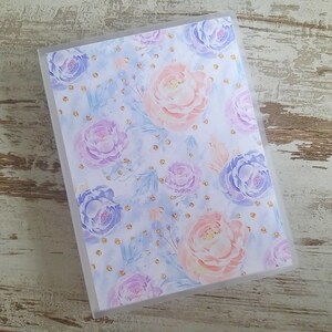 Puede incluir: Una carpeta de pl&aacute;stico transparente con un dise&ntilde;o floral. El dise&ntilde;o presenta flores rosas, moradas y azules con detalles de purpurina dorada sobre un fondo azul claro.