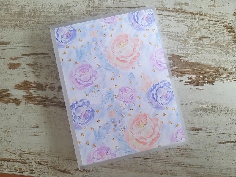 Puede incluir: Una carpeta de pl&aacute;stico transparente con un dise&ntilde;o floral. El dise&ntilde;o presenta flores rosas, moradas y azules con detalles de purpurina dorada sobre un fondo azul claro.
