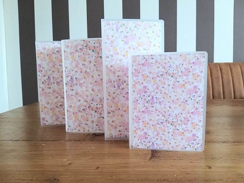 Puede incluir: Cinco carpetas de pl&aacute;stico transparente con un patr&oacute;n de confeti rosa y dorado. Las carpetas est&aacute;n apiladas sobre una mesa de madera.