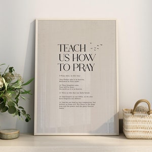 以下が含まれることがあります： ベージュの背景に黒い文字で「Teach Us How To Pray」と書いてある額装されたプリント。主の祈りが含まれています。プリントは、小さな白い籐のバスケット、白いカップ、本の束と一緒に棚の上に置かれています。