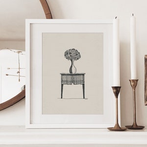 May include: A white framed print of a black and white line drawing of a vase with flowers on a small table. The print is hanging on a white wall above a white mantelpiece. Two candlesticks with white candles are on the mantelpiece, along with a small wooden frame with a black and white print inside.