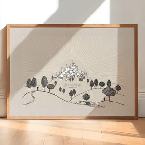 以下が含まれることがあります： 丘の上の町を白黒で描いたイラストが特徴の額入りアートプリント。木々や人物が描かれています。アートワークには、装飾的なフォントで「You are the light of the world...」というテキストが含まれています。フレームは明るい色の木材で作られています。
