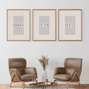 May include: Three framed prints with the text "SERMON ON THE MOUNT" in black text on a white background. The prints are hanging on a white wall above two brown leather armchairs and a coffee table with a vase of dried grass.