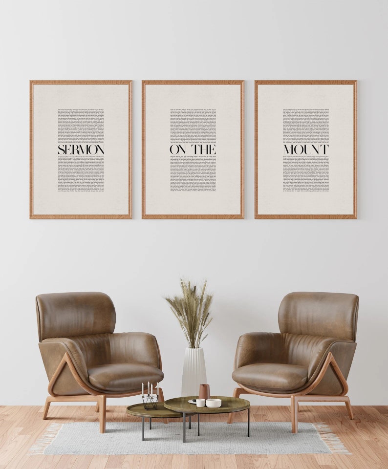 May include: Three framed prints with the text "SERMON ON THE MOUNT" in black text on a white background. The prints are hanging on a white wall above two brown leather armchairs and a coffee table with a vase of dried grass.