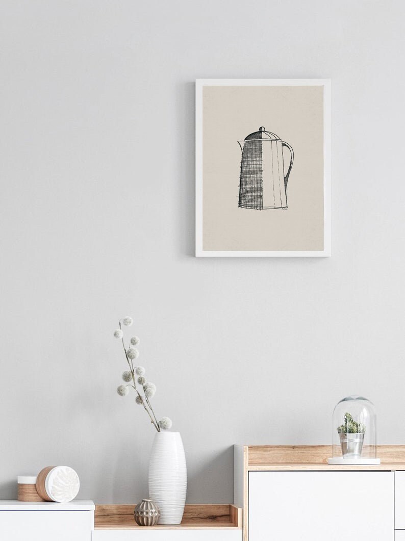 May include: A black and white line drawing of a coffee pot in a white frame on a light gray wall. The frame is hanging above a white dresser with a wooden top. A white vase with a dried flower arrangement is on the dresser.